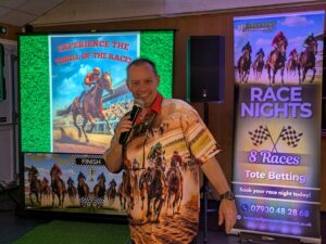 This photo is of Gary, the owner of Halfpint Entertainment. He's in a horse racing shirt standing in front of a screen with a horse racing image and a banner to his right stating Race Nights.